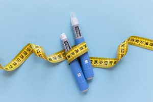Two injectors dosing pens for subcutaneous injection of antidiabetic medication or anti-obesity medication hovering over a blue background. Yellow measuring tape around the injectors.